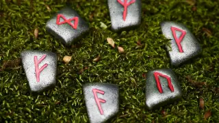 Kamienne runy z czerwonymi symbolami, w tym runa na pieniądze, leżą na zielonym mchu.