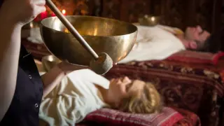 Terapia dźwiękiem z użyciem mis tybetańskich. Kobieta i mężczyzna leżą zrelaksowani, podczas gdy terapeuta uderza w misę.